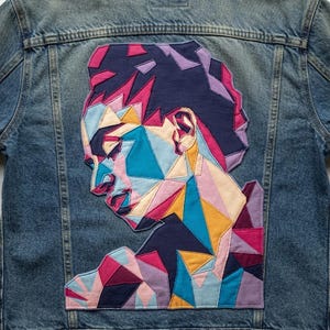 Könnte beinhalten: Eine blaue Jeansjacke mit einer großen, geometrischen Stoffapplikation auf der Rückseite. Das Design zeigt das Profil einer Person, das aus mehrfarbigen, kantigen Stoffstücken in Marineblau, Hellblau, Pink, Lila und Gelb zusammengesetzt ist.