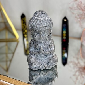 May include: A gray stone Buddha statue with a speckled finish, sitting in a meditative pose with hands clasped in front. The statue is on a reflective surface.