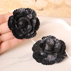 May include: Two black artificial flower blossoms with layered petals. The flowers are in full bloom, showcasing their intricate details. The blossoms are set against a neutral background, highlighting their dark colour and texture. These flowers could be used for crafting or decoration.