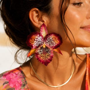 May include: A large statement earring shaped like an orchid flower, featuring deep burgundy and magenta petals with a white and black veined pattern, yellow accents at the center, and a smooth gold-toned chain necklace worn around the neck.