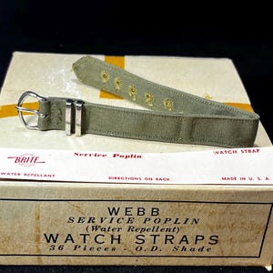Op de afbeelding: Een olijfgroene horlogeband met een zilveren gesp en decoratieve stiksels, liggend op een doos met het opschrift "Webb Service Poplin Watch Straps". De doos vermeldt ook "Water Repellent" en "Made in U.S.A."