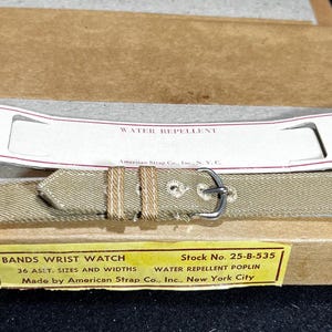 May include: A beige fabric watch band with a silver buckle. The band is packaged in a brown box with a yellow label that reads "Bands Wrist Watch, Stock No. 25-8-535, 36 Ast. Sizes and Widths, Water Repellent Poplin, Made by American Strap Co., Inc., New York City".