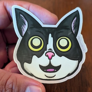 Può includere: Un adesivo raffigurante la faccia di un gatto dei cartoni animati con macchie bianche e nere, grandi occhi gialli e un naso rosa. Il gatto ha un'espressione sorridente. L'adesivo ha il testo "PROWORKS".