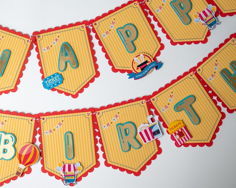 May include: A colorful banner with scalloped edges and red trim reads "Happy Birthday". Each letter is a different color and has a circus theme. The banner is decorated with small circus-themed images, including a ticket, popcorn, and a hot air balloon.