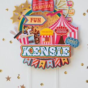May include: A colorful cake topper with a carnival theme. It features a Ferris wheel, a tent, a ticket booth, and the words "Welcome to the Carnival", "Fun", "Fantastic Circus", "Tickets", and "Kensile".
