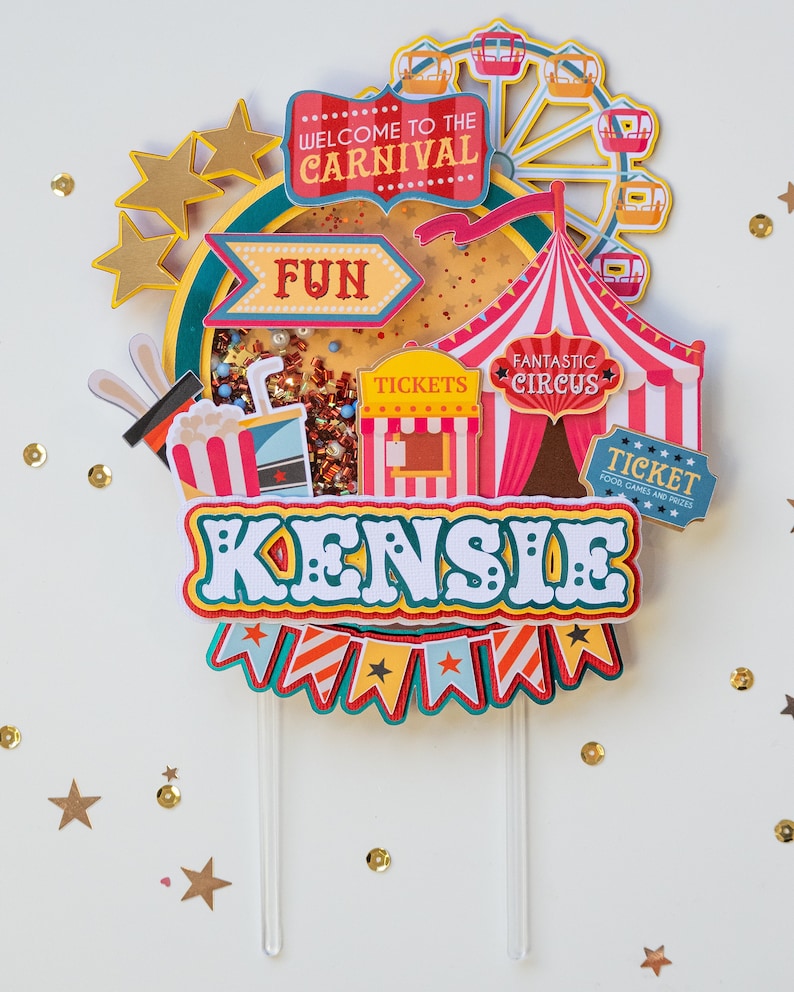 May include: A colorful cake topper with a carnival theme. It features a Ferris wheel, a tent, a ticket booth, and the words "Welcome to the Carnival", "Fun", "Fantastic Circus", "Tickets", and "Kensile".