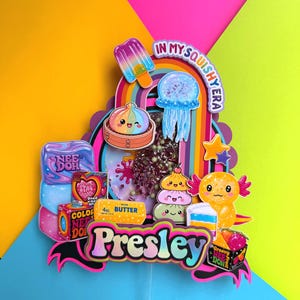Puede incluir: Un adorno para tarta multicapa con el nombre Presley en fuente negrita, rodeado de recortes coloridos de un polo, una medusa, un ajolote y varias cajas de juguetes. El centro incluye un elemento tipo shaker con cuentas y el texto In My Squishy Era.