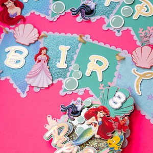 May include: A teal and white birthday banner with gold glitter and the words "Happy Birthday". The banner features Ariel, Ursula, and seashells. A separate topper features Ariel as a mermaid with the words "Happy Birthday" and the number "3" in a seashell.