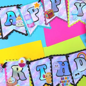 Puede incluir: Una guirnalda decorativa que deletrea HAPPY BIRTHDAY con banderines de estampado de galaxia morada, bordes festoneados y varios recortes tem&aacute;ticos de juguetes blandos, incluyendo un arco&iacute;ris, un cubo, una medusa y el texto IN MY SQUISHY ERA.