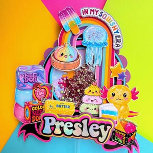 Puede incluir: Un adorno para pastel de varias capas con el nombre Presley en letras negritas, rodeado de recortes coloridos de un arco&iacute;ris, paleta, medusa, ajolote y varios paquetes de juguetes Nee Doh, con el texto In My Squishy Era en la parte superior.