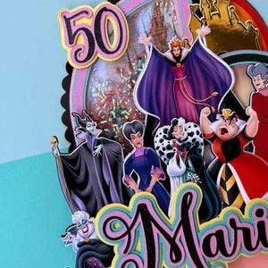 May include: A colorful cake topper featuring Disney villains, the number 50, and the name "Mari". The topper has a black scalloped edge, pink and blue accents, and a castle silhouette. The Evil Queen, Ursula, and Cruella de Vil are among the characters.