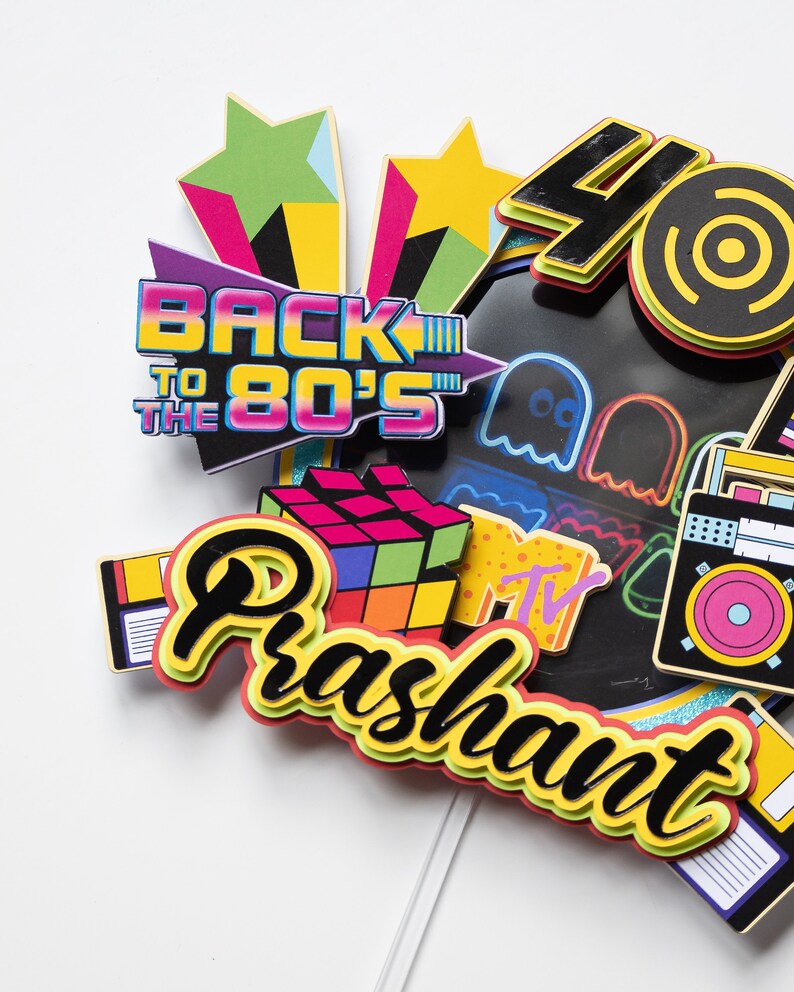 May include: A colorful cake topper with the words "Back to the 80's" and "Prashant" in a retro font. The topper features various 80's icons, including a Rubik's Cube, a cassette tape, a boombox, a Pac-Man ghost, and the MTV logo.