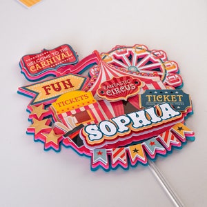 May include: A colorful cake topper with a circus theme. It features a red and white striped tent, a Ferris wheel, a ticket booth, and the words "Welcome to the Carnival", "Fun Tickets", "Fantastic Circus", "Ticket Food & Prizes", and "Sophia".