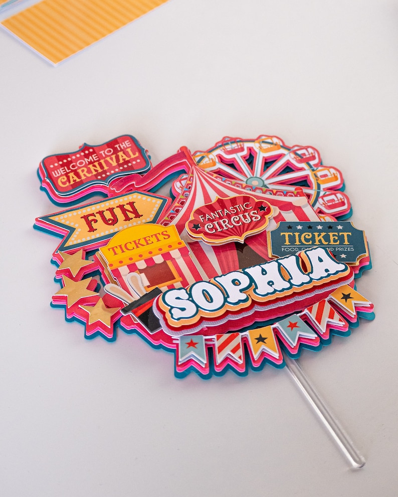 May include: A colorful cake topper with a circus theme. It features a red and white striped tent, a Ferris wheel, a ticket booth, and the words "Welcome to the Carnival", "Fun Tickets", "Fantastic Circus", "Ticket Food & Prizes", and "Sophia".