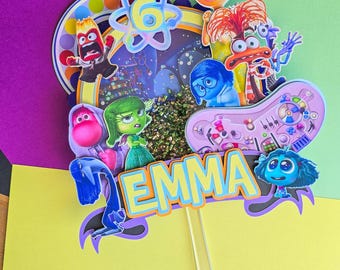 May include: A cake topper featuring characters from the movie Inside Out. The topper includes Joy, Sadness, Anger, Fear, and Disgust. The topper also includes the name "EMMA" and the number "6".