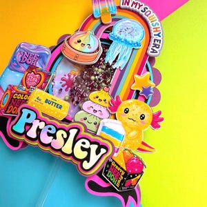 Puede incluir: Un adorno para pastel de varias capas con el nombre Presley en letras azules y amarillas. El dise&ntilde;o incluye gr&aacute;ficos coloridos de un helado, medusa, ajolote, dumplings y empaques de Nee Doh con el texto In My Squishy Era.