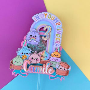 May include: A multi-layered cake topper featuring a central rainbow arch with the number 3 and the text IN MY DUMPLING ERA. The design includes various colorful, smiling dumpling characters in bamboo steamers and the name Camile in script.