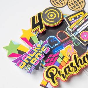 May include: A colorful cake topper with the text "Back to the 80s" and the number "40". The topper also features a Rubik's Cube, a cassette tape, a ghost, a star, and a disco ball. The name "Prasha" is written in a colorful font.