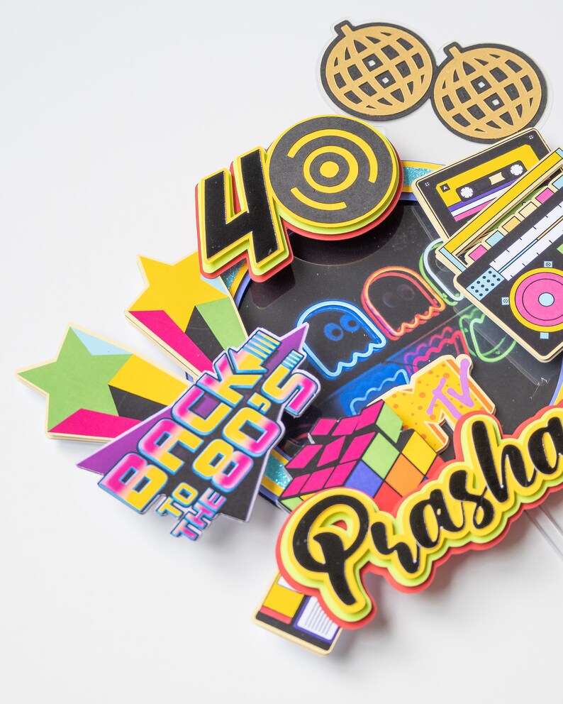 May include: A colorful cake topper with the text "Back to the 80s" and the number "40". The topper also features a Rubik's Cube, a cassette tape, a ghost, a star, and a disco ball. The name "Prasha" is written in a colorful font.