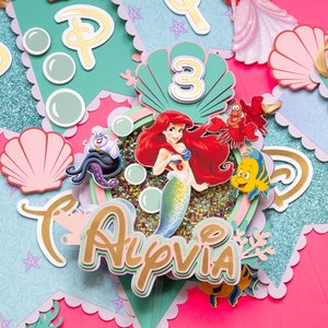 May include: A teal and gold banner with a mermaid, seashells, and the name "Alyvia" in gold letters. The banner also includes a number "3" and a trident.