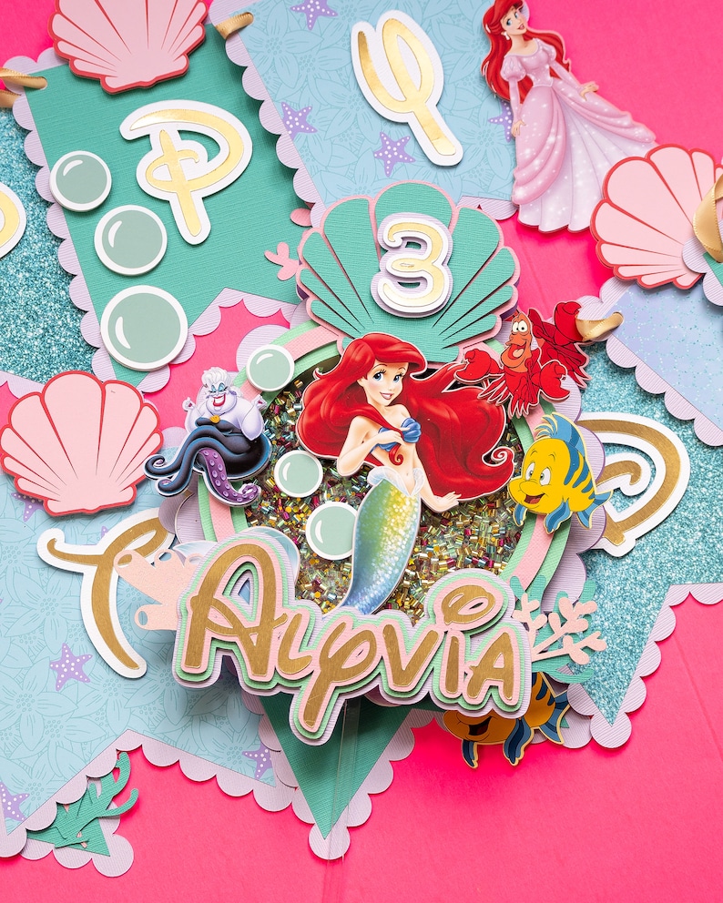 May include: A teal and gold banner with a mermaid, seashells, and the name "Alyvia" in gold letters. The banner also includes a number "3" and a trident.