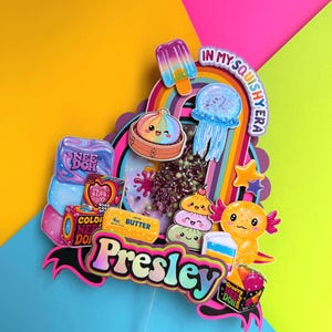 Puede incluir: Un adorno para pastel de varias capas con el nombre Presley en una fuente degradada, rodeado de recortes coloridos de una paleta, medusa, ajolote, una barra de mantequilla y varios juguetes, con el texto In My Squishy Era en la parte superior.