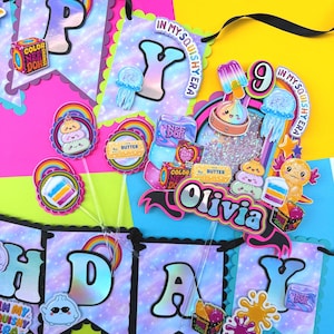 Puede incluir: Un conjunto de bander&iacute;n de cumplea&ntilde;os y adorno para pastel con fondo de galaxia y el texto "HAPPY" y "Olivia". Las decoraciones incluyen gr&aacute;ficos coloridos de juguetes, arco&iacute;ris, medusas y la marca "NEE DOH" sobre una cinta negra.