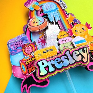 Puede incluir: Un adorno para pastel multicapa con el nombre Presley en letras negritas, rodeado de recortes coloridos que incluyen una medusa, un arco&iacute;ris, una barra de mantequilla de 113g y varias cajas de juguetes Nee Doh sobre un fondo multicolor.