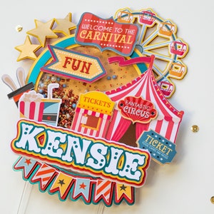 May include: A colorful carnival-themed cake topper with the name "KENSIE" in large, white letters. The topper features a Ferris wheel, a circus tent, a ticket booth, and a banner with stars and stripes.