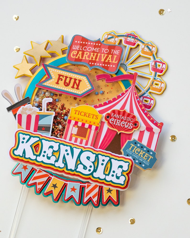 May include: A colorful carnival-themed cake topper with the name "KENSIE" in large, white letters. The topper features a Ferris wheel, a circus tent, a ticket booth, and a banner with stars and stripes.