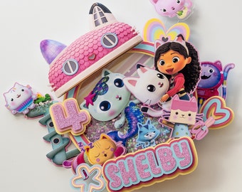 Peut inclure: Un topper de gâteau rose et bleu avec une maison de chat de dessin animé avec un arc-en-ciel et une fille aux cheveux bruns tenant un chat blanc. Le topper comprend également le chiffre 4 et le nom "Shelby" en lettres pailletées roses.