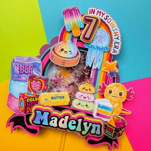 Puede incluir: Un adorno para tarta multicapa y colorido con el nombre Madelyn, el n&uacute;mero 7 y el texto In My Squishy Era. Incluye ilustraciones de un polo, medusa, ajolote y varios juguetes blandos, con un elemento central tipo coctelera lleno de cuentas.
