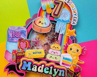 May include: A colorful layered cake topper featuring the name Madelyn, the number 7, and the text In My Squishy Era. It includes illustrations of a popsicle, jellyfish, axolotl, and various squishy toys, with a central shaker element filled with beads.