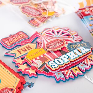 May include: A colorful set of circus-themed cake toppers with the words "Welcome to the Carnival", "Fun", "Tickets", "Fantastic Circus", "Ticket", and "Sophia".