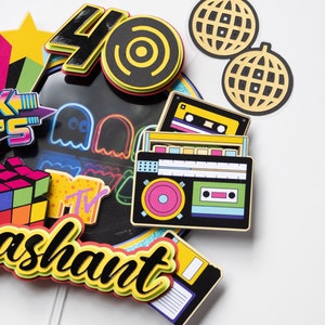May include: A colorful cake topper with a retro 80s theme. It features a number 40, a record, a cassette tape, a boombox, a Rubik's cube, a Pac-Man ghost, a disco ball, and the word "ashaunt".