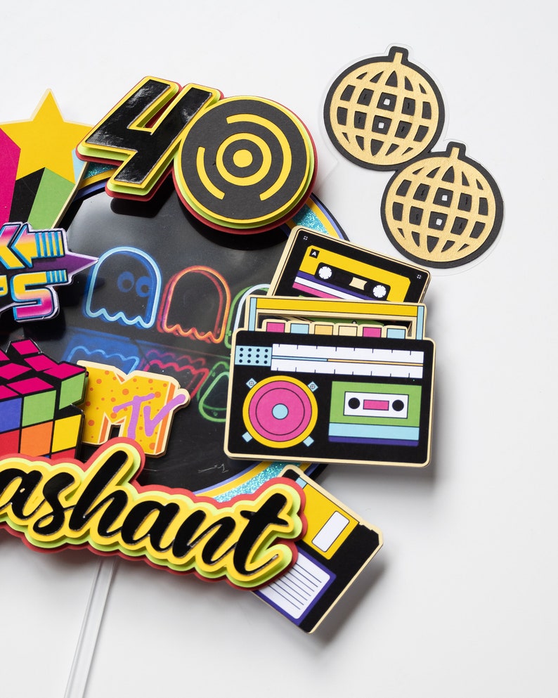 May include: A colorful cake topper with a retro 80s theme. It features a number 40, a record, a cassette tape, a boombox, a Rubik's cube, a Pac-Man ghost, a disco ball, and the word "ashaunt".