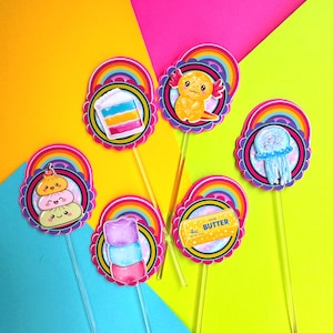 Puede incluir: Seis adornos decorativos para cupcakes sobre palitos de pl&aacute;stico transparente, cada uno con borde festoneado, un arco&iacute;ris y una ilustraci&oacute;n &uacute;nica que incluye un pastel en capas, un ajolote, una medusa, dumplings, malvaviscos y mantequilla.