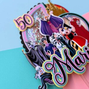 May include: A colorful cake topper featuring cartoon villains, the number 50, and the name "Mari". The topper includes images of Ursula, Maleficent, the Evil Queen, and Cruella de Vil. The design has a pink, blue, and black color scheme.