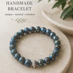 Könnte beinhalten: Ein handgefertigtes Armband mit schillernden blauen Perlen und einer silberfarbenen Akzentperle. Das Armband wird auf einer neutralen Oberfläche präsentiert. Der Text "HANDMADE BRACELET" steht oben, darunter "unique • natural • timeless".