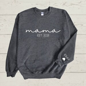 May include: Dark grey sweatshirt with the word "mama" in white script, and "EST. 2021" below it. The sleeve features the name "Piper" and a small white heart. The sweatshirt is made of a soft material.