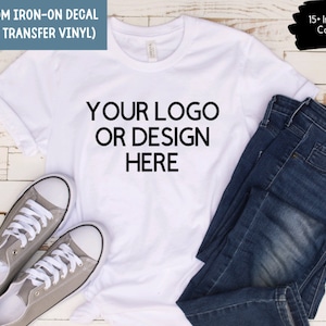 Może przedstawiać: Biała koszulka z nadrukiem "YOUR LOGO OR DESIGN HERE" w kolorze czarnym. Koszulka leży płasko na drewnianej powierzchni z parą szarych butów sportowych i niebieskimi dżinsami.