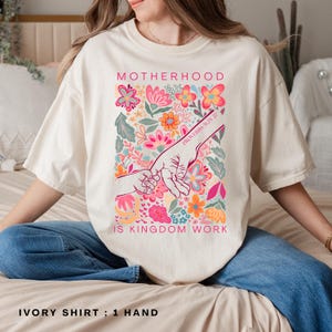 May include: Ivory-colored t-shirt with the words "MOTHERHOOD IS KINGDOM WORK" in pink. The shirt features a floral design with a hand-holding graphic. The text "PROVERBS 31:25" is also visible.