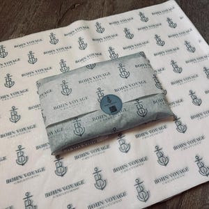 May include: A package wrapped in gray tissue paper with a repeating pattern of anchors and the text "Bohn Voyage Creations." A round sticker seals the package. The tissue paper is laid on a larger sheet with the same design.