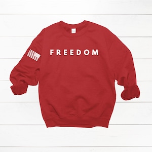 May include: A red sweatshirt with the word "FREEDOM" printed in white across the chest. The left sleeve features a small American flag. The sweatshirt is displayed on a white wooden surface.