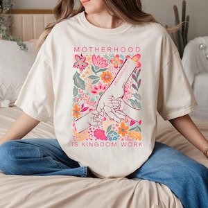 May include: Cream-colored t-shirt with a floral design and the text "MOTHERHOOD IS KINGDOM WORK." The design includes hands holding each other, with the words "PROVERBS 31:25-27" on one hand. The floral arrangement features pink, orange, and blue flowers.