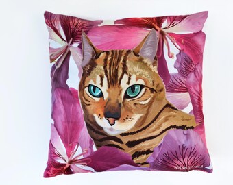 Peut inclure: Un coussin carré représentant un chat tigré brun aux yeux bleu sarcelle saisissants, centré sur un fond vibrant de grandes fleurs rose foncé et violettes rappelant des orchidées. Le nom Namoshka est imprimé en petites lettres blanches en bas.
