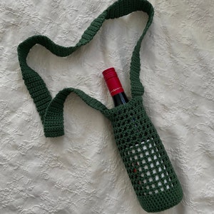 May include: A green crocheted wine bottle carrier with a long strap. The carrier is made of a mesh pattern and has a bottle of wine inside.