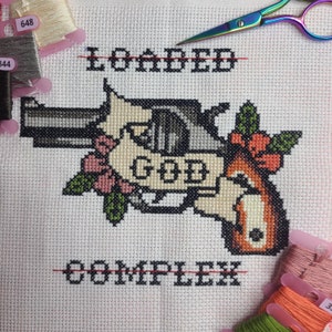 Könnte beinhalten: Kreuzstich-Stickerei einer Pistole mit einer Blume und den Worten "LOADED" und "COMPLEX" darüber und darunter. Die Pistole trägt das Wort "GOD" auf sich.