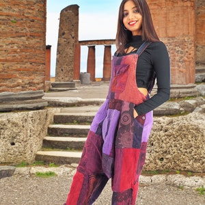 May include: A woman wearing a patchwork jumpsuit with a purple, red, and black color scheme. She is standing in front of a stone structure with columns.