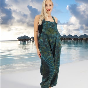 May include: A woman wearing a green and black tie-dye jumpsuit with a single pocket on the front. She is standing on a white sandy beach with blue water in the background.